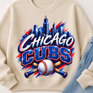 Puede incluir: Una sudadera beige de cuello redondo con un estampado gráfico del horizonte de Chicago en azul y blanco, con el texto CHICAGO CUBS en letras azules en negrita, dos bates de béisbol cruzados y una pelota de béisbol blanca con costuras rojas.