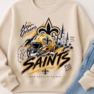 Op de afbeelding: Een beige sweater met ronde hals en een grafische print van een gouden footballhelm, een fleur-de-lis en een kathedraal. De tekst luidt New Orleans, Saints, Who Dat, Black & Gold Forever en New Orleans Saints met een spelschema.