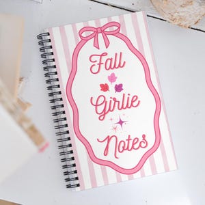 Puede incluir: Un cuaderno de espiral con una cubierta de rayas verticales blancas y rosa claro. El centro presenta una forma delineada en rosa que recuerda a una etiqueta con lazo, con el texto Fall Girlie Notes, tres pequeñas hojas coloridas y dos estrellas.