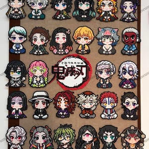 May include: A collection of twenty-nine pixel art figures arranged on a brown board, featuring various anime characters with distinct hair colours and outfits. A central circular logo displays the Japanese text 鬼滅の刃 in black on a white background.