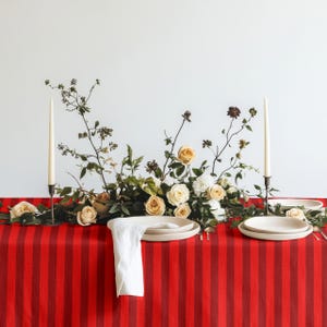 May include: A table setting featuring a red and black striped tablecloth, two tall white taper candles in dark holders, and a central floral arrangement of cream and white roses with green foliage. Two white plates and a white napkin are included.