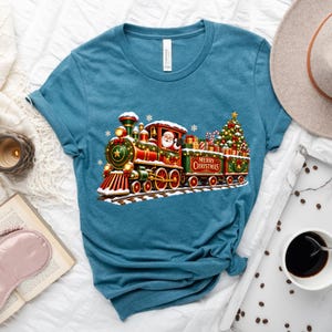 May include: A teal blue t-shirt featuring a graphic of a festive steam train decorated with Christmas lights, a star, and a wreath. Santa Claus is visible in the engine cab, and the train cars carry wrapped gifts and a Christmas tree.