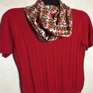 Puede incluir: Un suéter rojo de punto con mangas cortas y una bufanda colorida con un patrón geométrico alrededor del cuello.