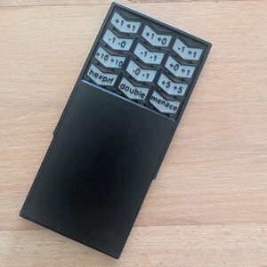 Op de afbeelding: Een zwart rechthoekig spelaccessoire met een schuifpaneel dat een raster van lichtgrijze, chevronvormige knoppen onthult. De knoppen zijn voorzien van diverse numerieke waarden en de tekst hexproof, double en menace in een zwart lettertype.