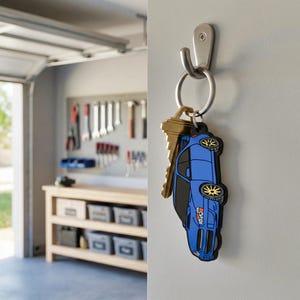 May include: A metal key ring hangs from a silver wall hook, holding a brass key and a blue car-shaped keychain. The keychain features a detailed blue sedan with black windows, yellow-rimmed wheels, and a small orange graphic on the front fender.