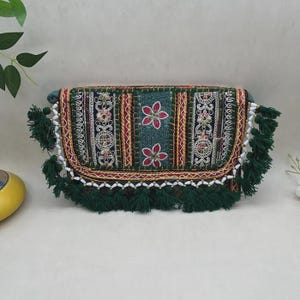 May include: A rectangular clutch bag featuring a dark green fabric base with intricate embroidery in red, gold, and white. The flap is decorated with floral patterns and geometric borders, finished with dark green tassels and white beads along the edge.