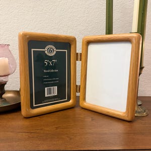 May include: A hinged double picture frame made of light-colored wood with rounded edges. The left frame contains a dark green insert with the text Windsor & Browne, 5"x7", Wood Collection, and a barcode. The right frame is empty with a white backing.