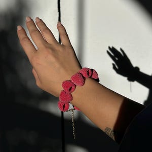 Może przedstawiać: Bransoletka z rzędem teksturowanych zawieszek w kształcie malin w kolorze ciemnego różu. Zawieszki są połączone cienkim, złotym metalowym łańcuszkiem, który zawiera małą regulowaną przedłużkę z zapięciem typu karabińczyk.