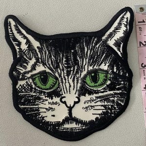 Puede incluir: Un parche bordado de cabeza de gato con detalles de hilo negro y blanco y ojos de color verde brillante. El parche está colocado junto a una cinta métrica rosa, que muestra que la altura de la cabeza del gato es de unos 11,5 cm.