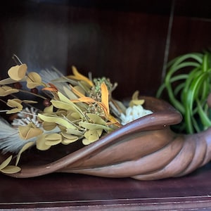 May include: A brown ceramic cornucopia vessel filled with an arrangement of dried botanical elements, including pale yellow leaves, white feathery plumes, and small dark berries, creating a textured display of natural autumn-toned foliage.