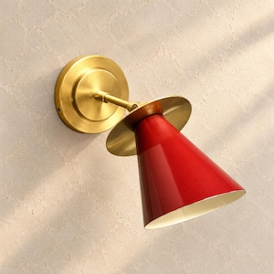 Peut inclure: Une applique murale composée d'une base circulaire en laiton brossé et d'un bras assorti. La tête de la lampe est un abat-jour conique rouge vif doté d'un large disque en laiton, offrant une source lumineuse ciblée avec une finition métallique.