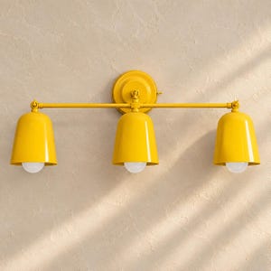 Peut inclure: Une applique murale jaune vif composée d'une barre horizontale et de trois abat-jours en forme de cloche orientés vers le bas. Chaque abat-jour contient une ampoule blanche, et l'ensemble est fixé à une plaque de base circulaire centrale.