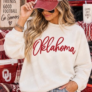 Op de afbeelding: Een crèmekleurige sweater met ronde hals en het woord Oklahoma in donkerrode cursieve letters op de borst gedrukt. Het kledingstuk heeft lange mouwen en een relaxte pasvorm, gemaakt van zachte stof met een geribbelde halslijn en boorden.