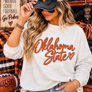 Op de afbeelding: Een wit sweatshirt met lange mouwen en de oranje tekst Oklahoma State met een hartje. Het kledingstuk wordt gecombineerd met een zwarte honkbalpet met de tekst GAME DAY en een hartje, plus een blauwe spijkerbroek en kralenarmbanden.