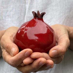 May include: A glossy, deep red ceramic pomegranate sculpture with a dark brown, textured calyx at the top. The surface of the fruit is smooth and reflective, showcasing a rounded, bulbous shape that mimics the appearance of a real pomegranate.