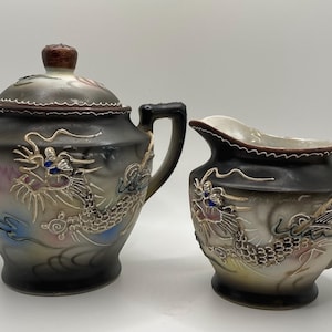 May include: A ceramic sugar bowl with a lid and a matching creamer pitcher. Both pieces feature a dark brown and cream gradient finish with a raised, textured dragon motif in gold, blue, and white tones, complete with two handles on the sugar bowl.
