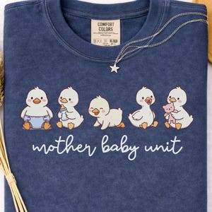 May include: A navy blue crewneck t-shirt featuring a graphic of five white ducklings in various poses, including one with a diaper, a bottle, a pacifier, and a teddy bear. Below the ducks, the text reads mother baby unit in white script.
