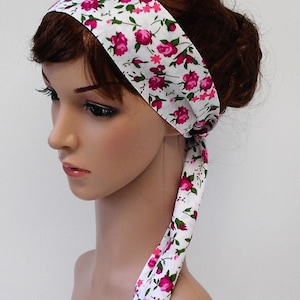 May include: A white headband with a floral pattern of pink roses and green leaves. The headband is tied at the back, with the ends hanging down. The accessory is designed to be worn on the head.
