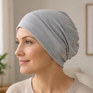May include: A light grey beanie hat made of a soft, jersey-knit fabric. The hat features a relaxed, slouchy fit that covers the head completely, with subtle folds and gathers at the back, providing a smooth and comfortable appearance.