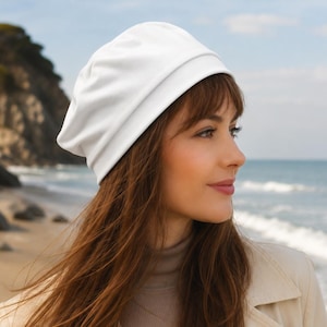 May include: A solid white slouchy beanie hat made of a smooth, lightweight fabric. The beanie features a rounded crown and a folded brim edge, designed to fit loosely around the head with a soft, draped silhouette that sits comfortably above the ears.
