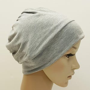 May include: A light gray beanie hat made of soft fabric. The hat is shown on a mannequin head, with the fabric gathered at the top and a folded brim. The hat is a solid color and appears to be made of a knit material.