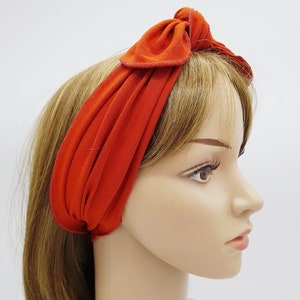 Pode incluir: Uma faixa de cabelo de tecido laranja brilhante com um laço amarrado no topo. A faixa de cabelo é usada em um manequim com cabelo castanho longo.