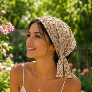 May include: A triangular head scarf featuring a small-scale floral pattern with pink, green, and white blossoms on a light beige background. The fabric is tied at the back with two long, pointed ends, creating a classic bandana-style hair accessory.