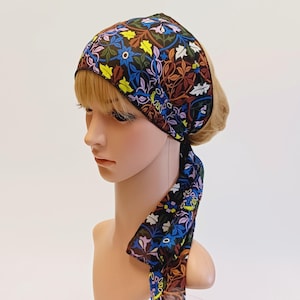 Könnte beinhalten: Ein gemustertes Kopftuch mit einem floralen Design in Blau-, Gelb-, Braun- und Weißtönen auf schwarzem Hintergrund. Das Tuch ist hinten gebunden, die Enden hängen herunter. Die Kopfbedeckung wird auf einem Schaufensterkopf getragen.