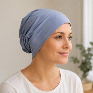 May include: A solid periwinkle blue slouchy beanie hat made of a smooth, soft fabric. The hat features a wide, fitted band around the forehead and a relaxed, gathered crown that creates a draped, voluminous silhouette at the back of the head.