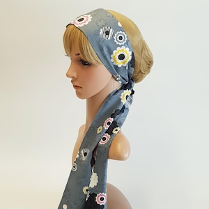 Puede incluir: Una diadema gris con un estampado floral con flores blancas, amarillas, rosas y negras. La diadema está atada en la parte posterior y cae hacia abajo. La tela tiene una textura sutil.