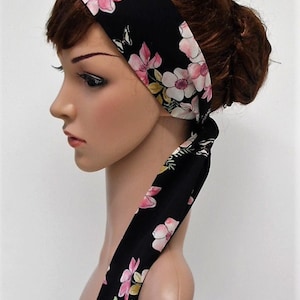 May include: Black floral headband with pink and white flowers. The headband is tied at the back, with long, flowing fabric. The floral pattern includes green leaves. A stylish hair accessory for any occasion.