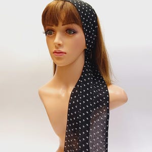 May include: A black headscarf with white polka dots is draped over a mannequin head. The scarf is tied at the top of the head and hangs down the back. The mannequin has brown hair and is set against a white background.