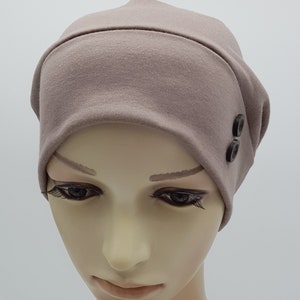 May include: A soft, light brown beanie with two black buttons on the side. The beanie is made of a stretchy fabric and is designed to fit comfortably on the head.