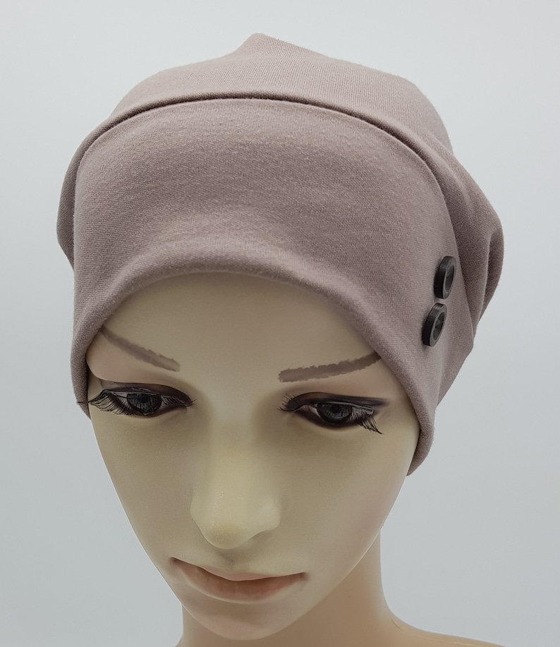 May include: A soft, light brown beanie with two black buttons on the side. The beanie is made of a stretchy fabric and is designed to fit comfortably on the head.