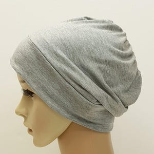 May include: A light gray beanie hat made of soft fabric. The hat is shown on a mannequin head, with the fabric draped and gathered at the back. The beanie is a solid color and appears to be a comfortable, casual accessory.