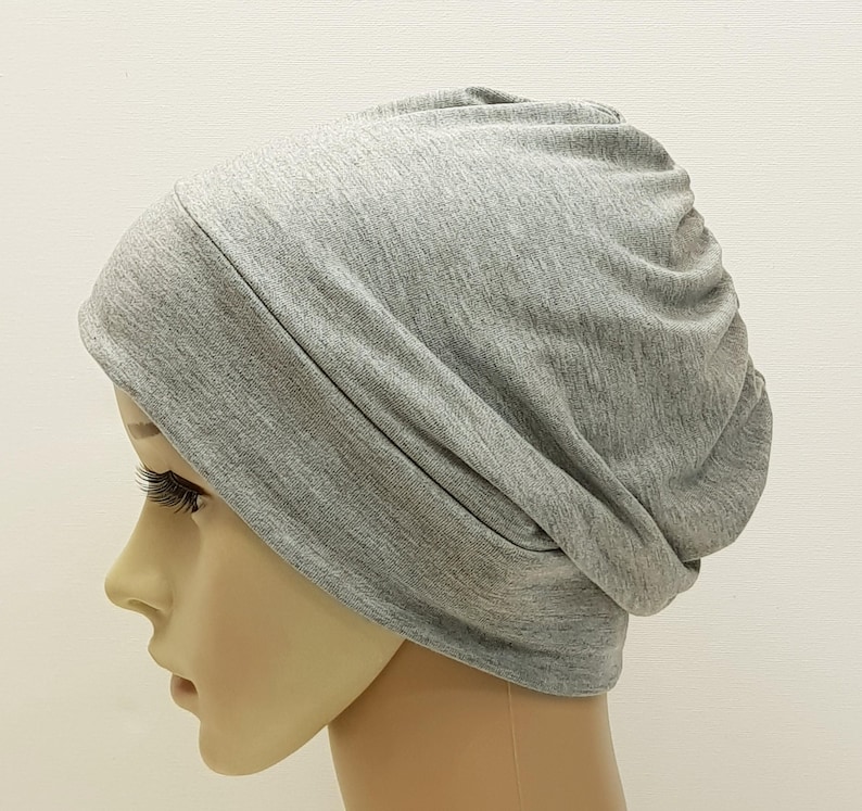 May include: A light gray beanie hat made of soft fabric. The hat is shown on a mannequin head, with the fabric draped and gathered at the back. The beanie is a solid color and appears to be a comfortable, casual accessory.