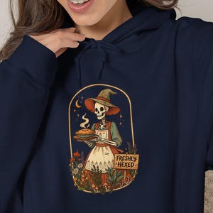 May include: A navy blue hooded sweatshirt featuring a graphic print of a skeleton wearing a witch hat and apron, holding a steaming pie. The design includes flowers, mushrooms, and a wooden sign with the text FRESHLY HEXED in capital letters.