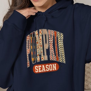 May include: A navy blue hooded sweatshirt featuring the text PUMPKIN SEASON in a bold, arched font. The letters of PUMPKIN are filled with various autumn-themed patterns including plaid, stripes, and floral motifs in orange, red, and white.