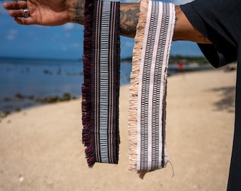 May include: Two rectangular woven fabric strips featuring intricate geometric patterns in black, white, and maroon. Each strip is finished with long, frayed fringe edges along one side, showcasing a textured, multi-layered textile design.
