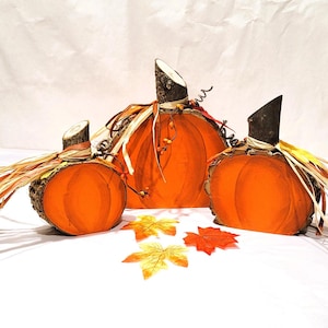 May include: Three orange pumpkin decorations with wooden stems and raffia accents. The pumpkins vary in size and are arranged with colorful fall leaves, creating a festive autumn display. The pumpkins are made of fabric.