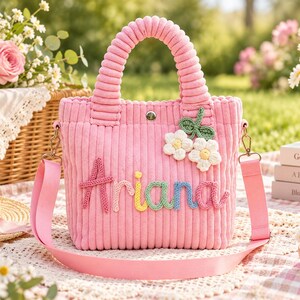 May include: A pink corduroy tote bag featuring the name Ariana in multi-colored yarn lettering. The bag has two top handles, a detachable shoulder strap, a silver snap closure, and two white crocheted flower appliqués with green leaves.
