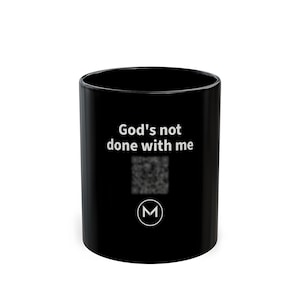 May include: A black ceramic mug featuring the white text "God's not done with me" printed on the front. Below the text is a blurred square QR code and a white circular logo containing the letter M in a stylized font.