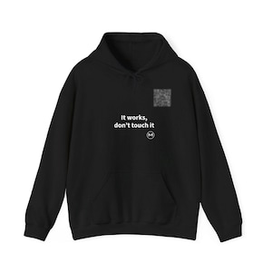 May include: A black hooded sweatshirt featuring a front kangaroo pocket and long sleeves. The chest area displays the white text "It works, don't touch it" positioned above a small circular logo with the letter M inside.
