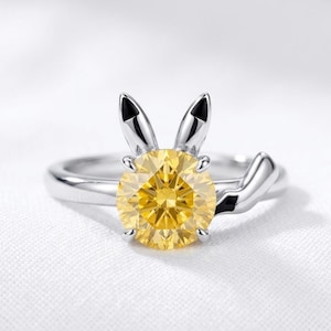 Puede incluir: Un anillo de tono plateado con una piedra preciosa amarilla redonda de talla brillante en el centro. El engaste presenta dos orejas plateadas puntiagudas hacia arriba y un elemento de cola plateada curvada en el lateral del anillo.