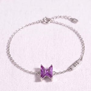 May include: A silver-toned chain bracelet featuring a central purple, butterfly-shaped gemstone and a small rectangular metal tag engraved with the letters BTS. The delicate link chain includes a circular clasp and a small jump ring for closure.
