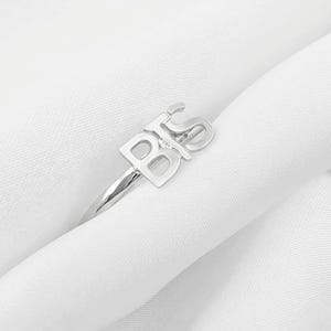 Puede incluir: Un anillo de metal en tono plateado que presenta las letras BTS como punto focal. Las letras están conectadas en un diseño continuo y pulido que se asienta sobre una banda delgada y lisa. El anillo tiene un acabado metálico reflectante.