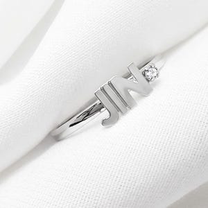 Puede incluir: Un anillo de metal plateado que presenta las letras JIN en una fuente mayúscula negrita integrada en la banda. Una pequeña piedra preciosa redonda y transparente está engastada en la banda junto a la letra N, añadiendo un brillo sutil.