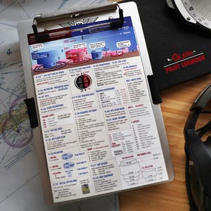May include: A silver metal clipboard holding a white reference sheet with blue, red, and black text and diagrams detailing aviation regulations, airspace classifications, V-speeds, and flight requirements, positioned next to a black pilot logbook.
