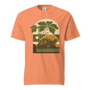 以下が含まれることがあります： ピーチカラーの半袖Tシャツ。白い花をつけた大きな緑の植物の下を歩くカメのグラフィックプリントが施されています。デザインはアーチ型のフレームの中にあり、背景には黄色い太陽と水平線が描かれています。