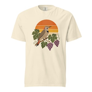 May include: A cream-colored short-sleeve t-shirt featuring a graphic print of a northern flicker bird perched on a grapevine branch with purple grapes and green leaves, set against a circular orange and yellow sunset background design.